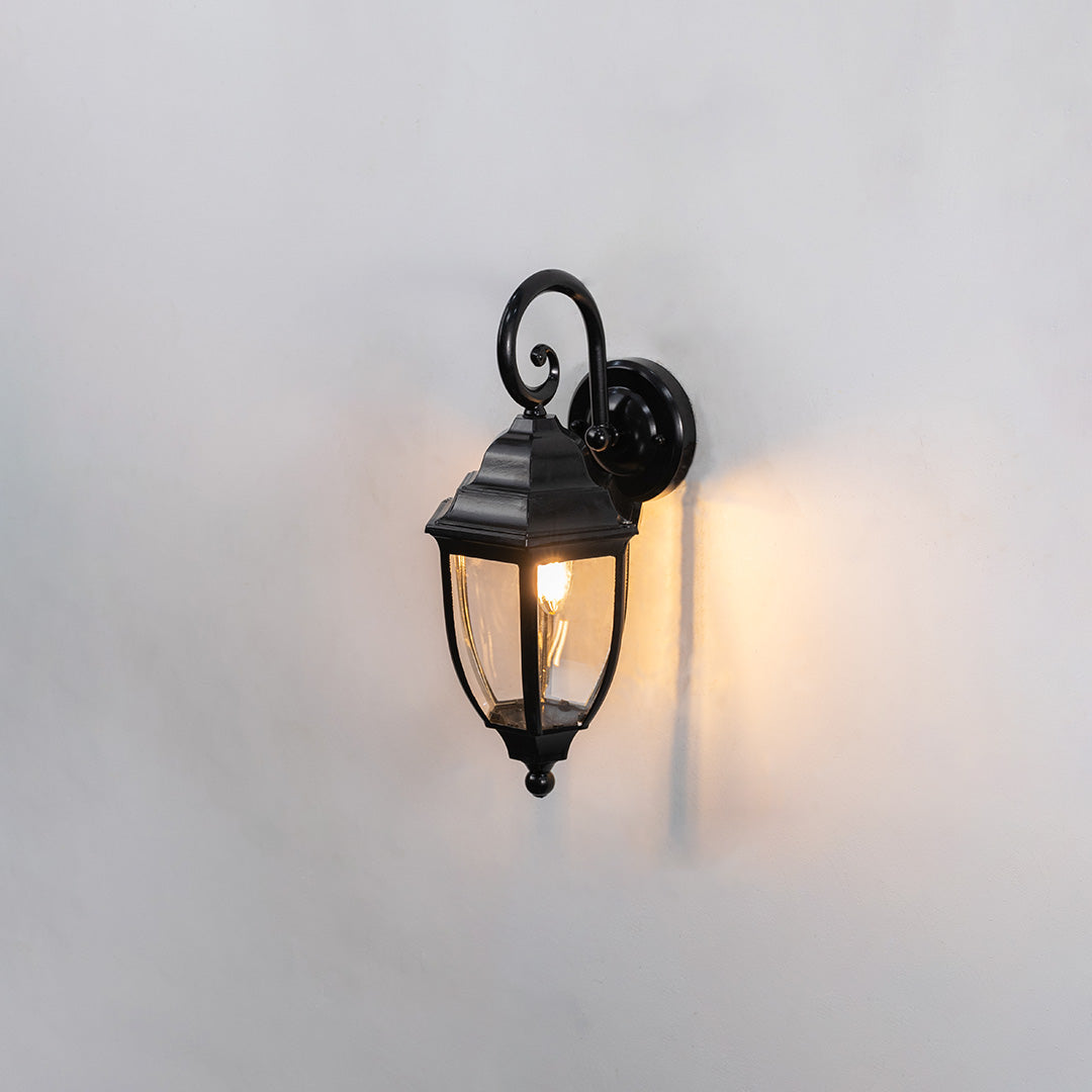Black metal outdoor wall light on white background with a classic glass lantern shade casting a soft warm glow for decor.