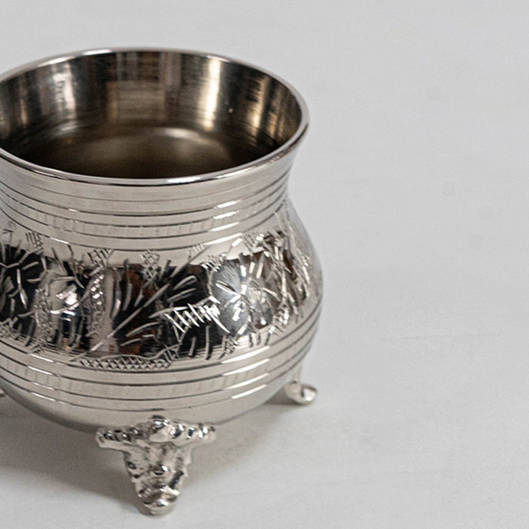 Closeup of silver teapot with ornate detailing curved spout lid and handle on footed base placed on white background.