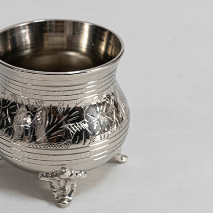 Closeup of silver teapot with ornate detailing curved spout lid and handle on footed base placed on white background.