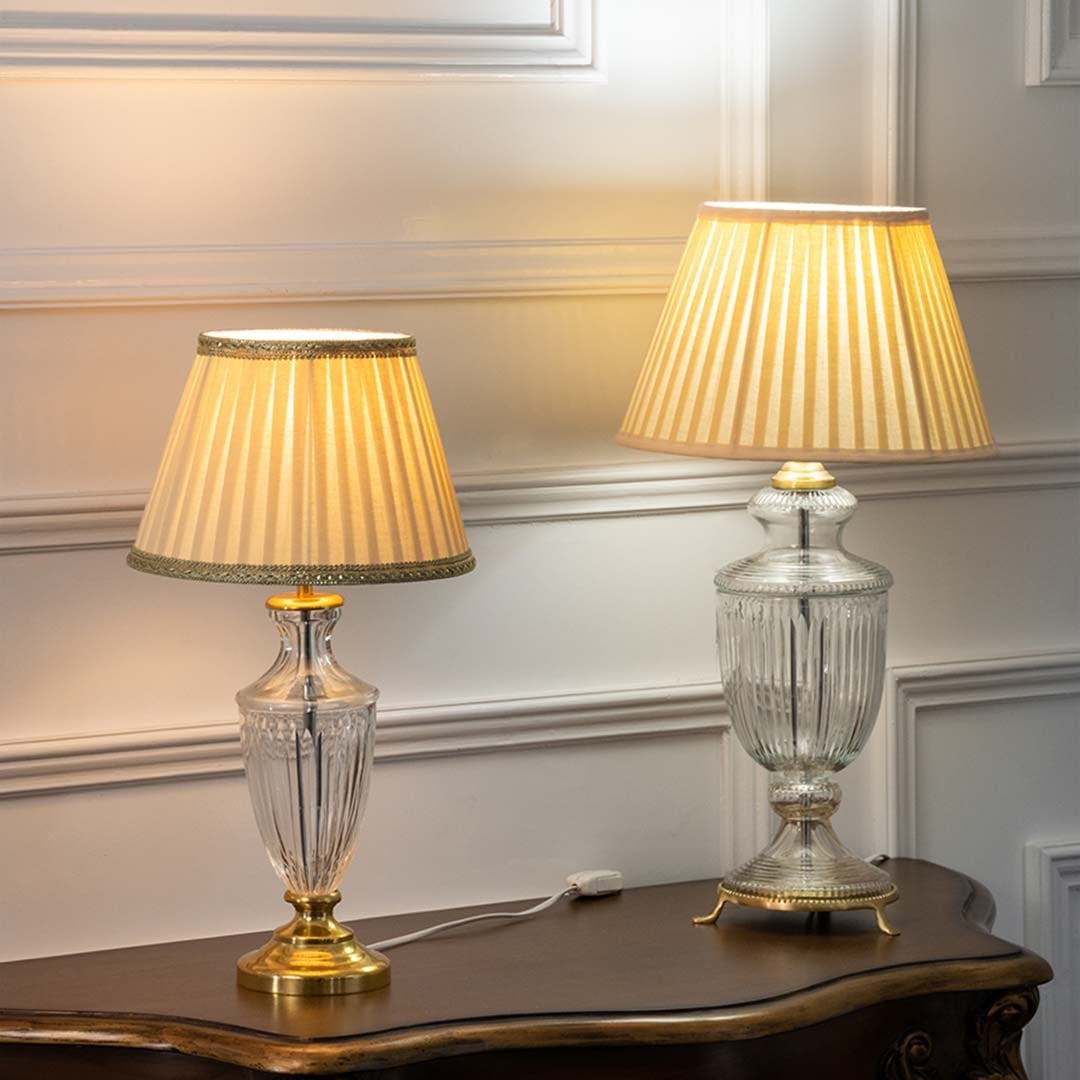 Pair of crystal glass table lamps with gold bases and pleated beige shades turned on together adding soft warm illumination to classic home interiors.