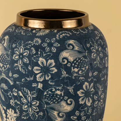 Closeup image of a blue ceramic decor vase with white floral pattern and metallic rim shown clearly on a soft light background highlighting surface texture and detailing.