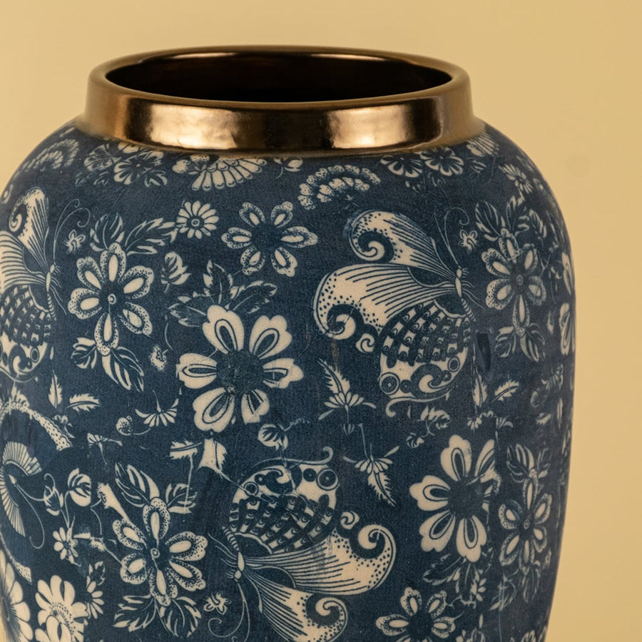 Closeup image of a blue ceramic decor vase with white floral pattern and metallic rim shown clearly on a soft light background highlighting surface texture and detailing.
