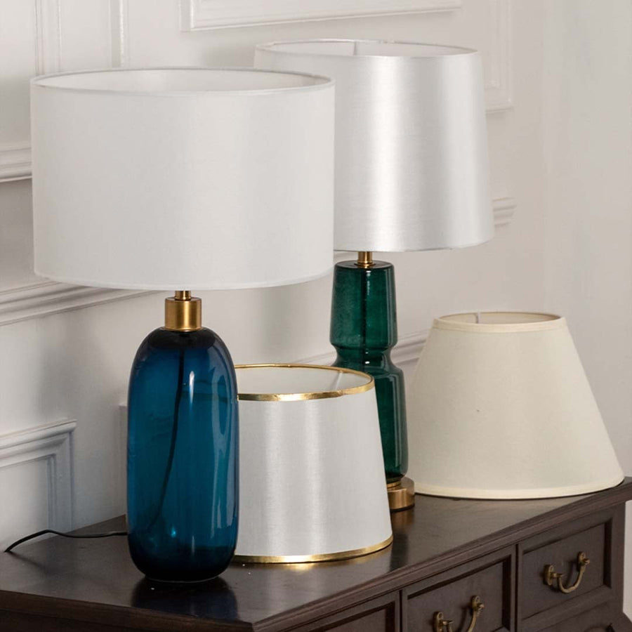 Teal glass table lamp with white shade on wooden sideboard with light off for fresh modern décor styling.