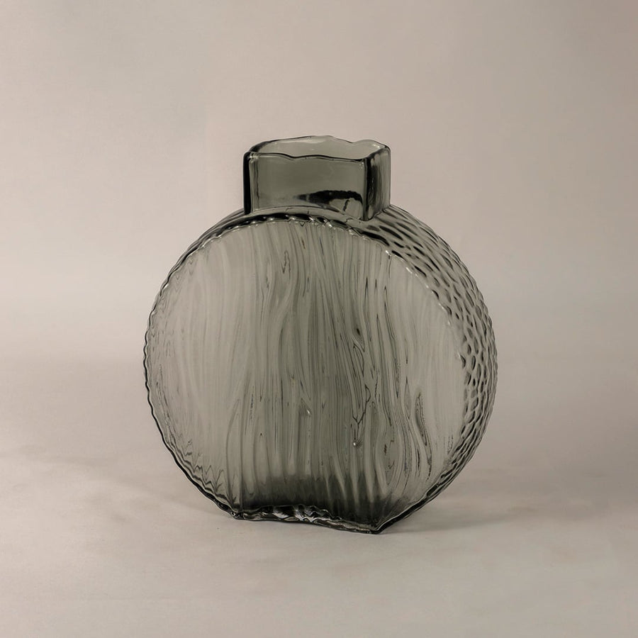 Side angle of round smoky gray glass vase showing its curved form  ribbed texture  and smooth reflective surface.