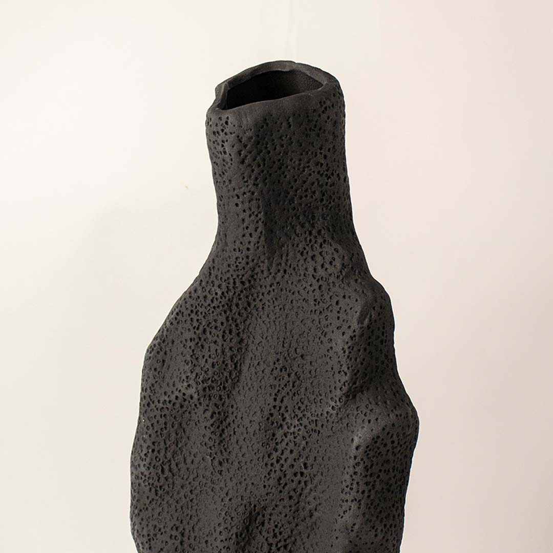 Dark textured ceramic vase closeup with organic sculptural shape placed on white surface against neutral background showcasing rustic modern home decor styling.