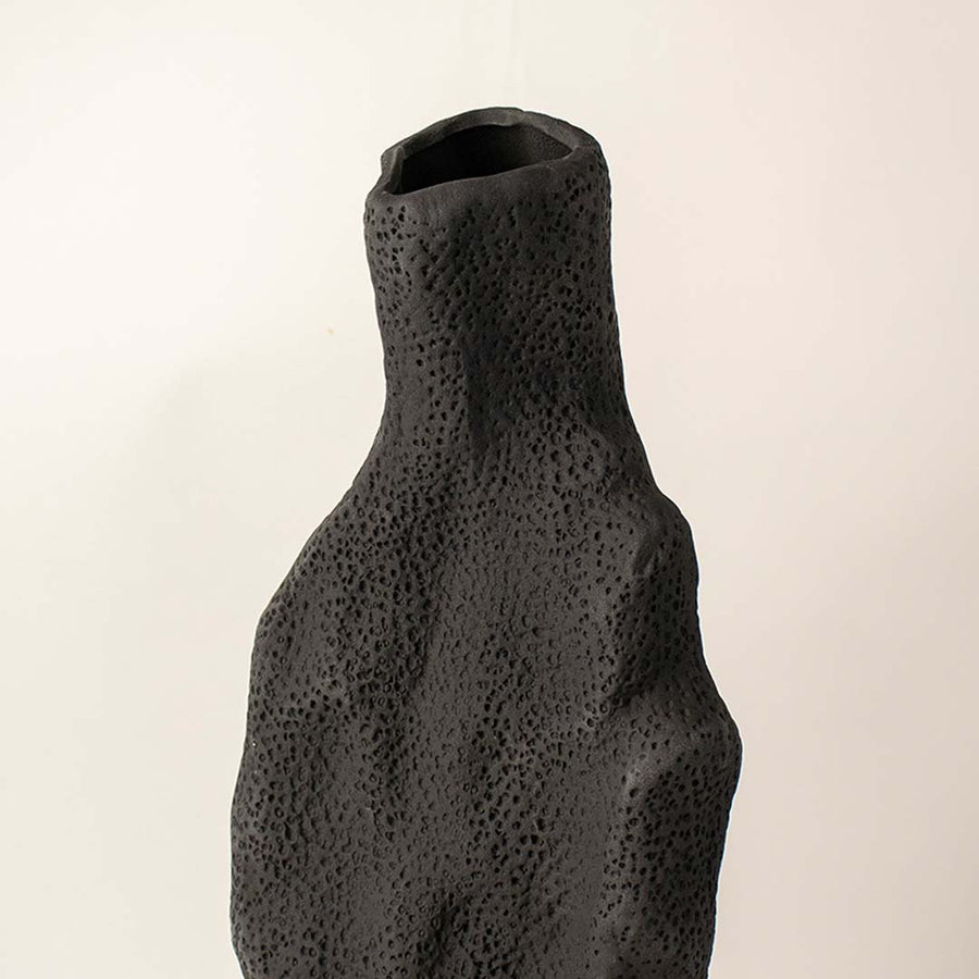 Dark textured ceramic vase closeup with organic sculptural shape placed on white surface against neutral background showcasing rustic modern home decor styling.
