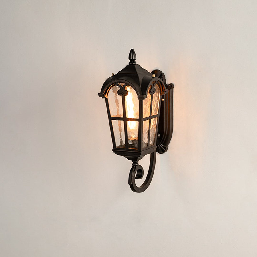 Black metal outdoor wall light on white background with a textured water glass shade and ornate bottom scroll design.