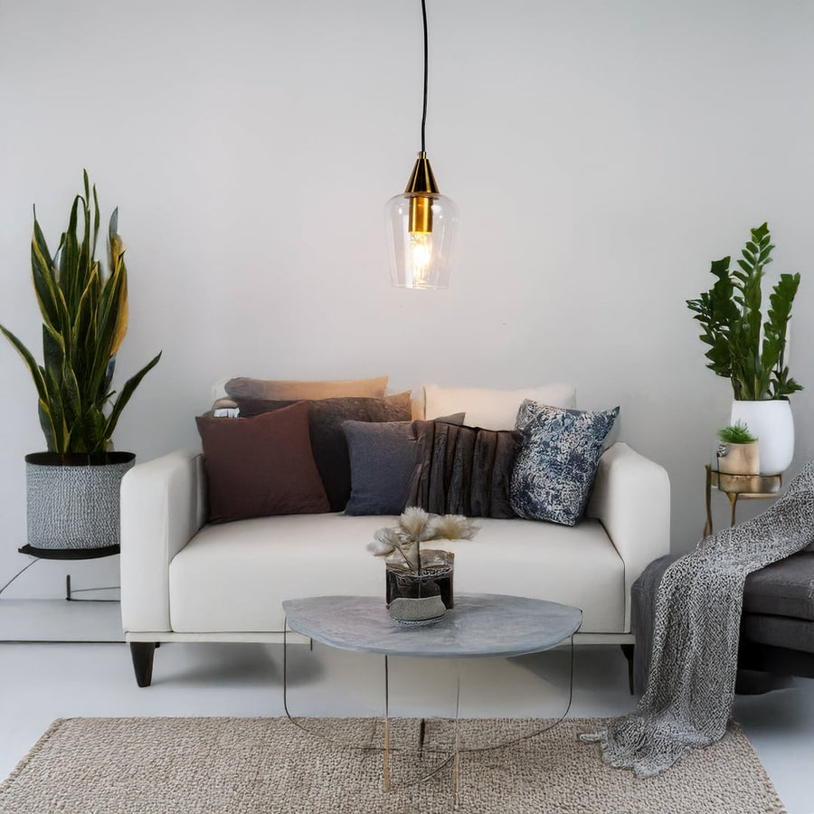 A modern glass pendant light with a gold top illuminating a white sofa with mixed cushions and plants creating a soft stylish living room setting.
