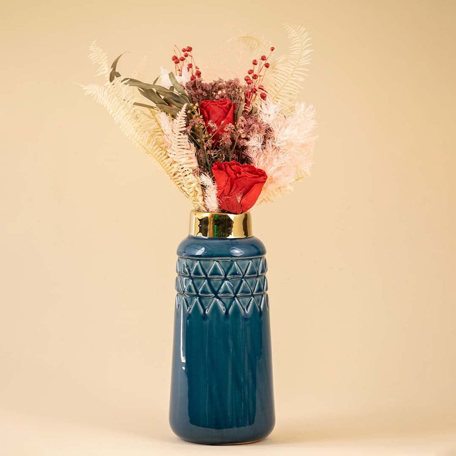 Elegant dark blue ceramic vase with a gold rim filled with red roses ferns and dried flowers placed against a warm beige background creating a rich and luxurious decorative look.