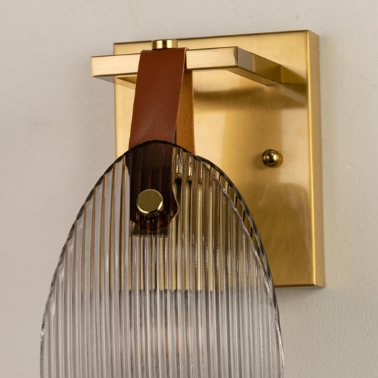 Closeup of gold metal wall light base with ribbed glass shade and leather strap accent on white background for premium interior decor.