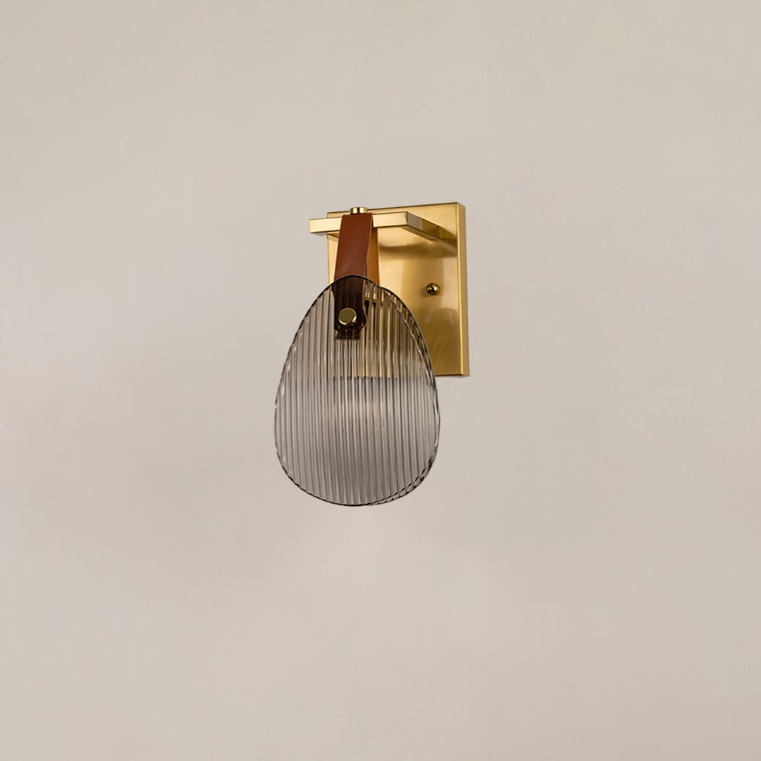 Gold metal wall sconce with ribbed glass and leather strap illuminated on white background for elegant lighting.
