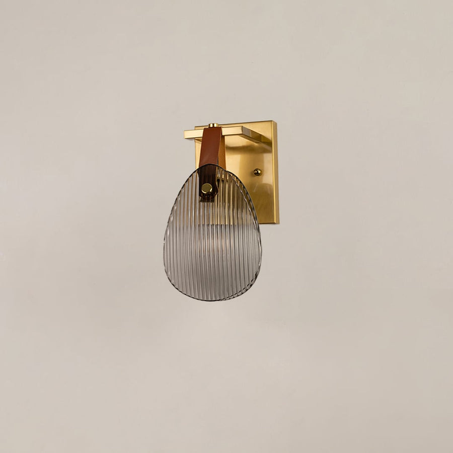 Gold metal wall sconce with ribbed glass and leather strap illuminated on white background for elegant lighting.