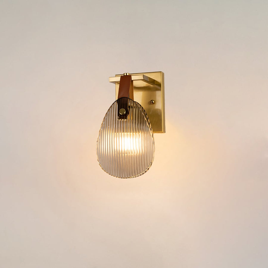 Gold wall light with ribbed glass shade and leather strap glowing on a white background for modern home lighting.