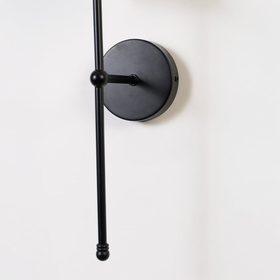 Closeup of black wall light showing the slim metal rod and white shade with a modern minimal finish.