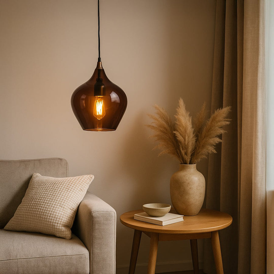 A warm amber glass pendant light glowing softly above a neutral living room setup with a beige sofa wooden side table and pampas decor creating a cozy ambiance.