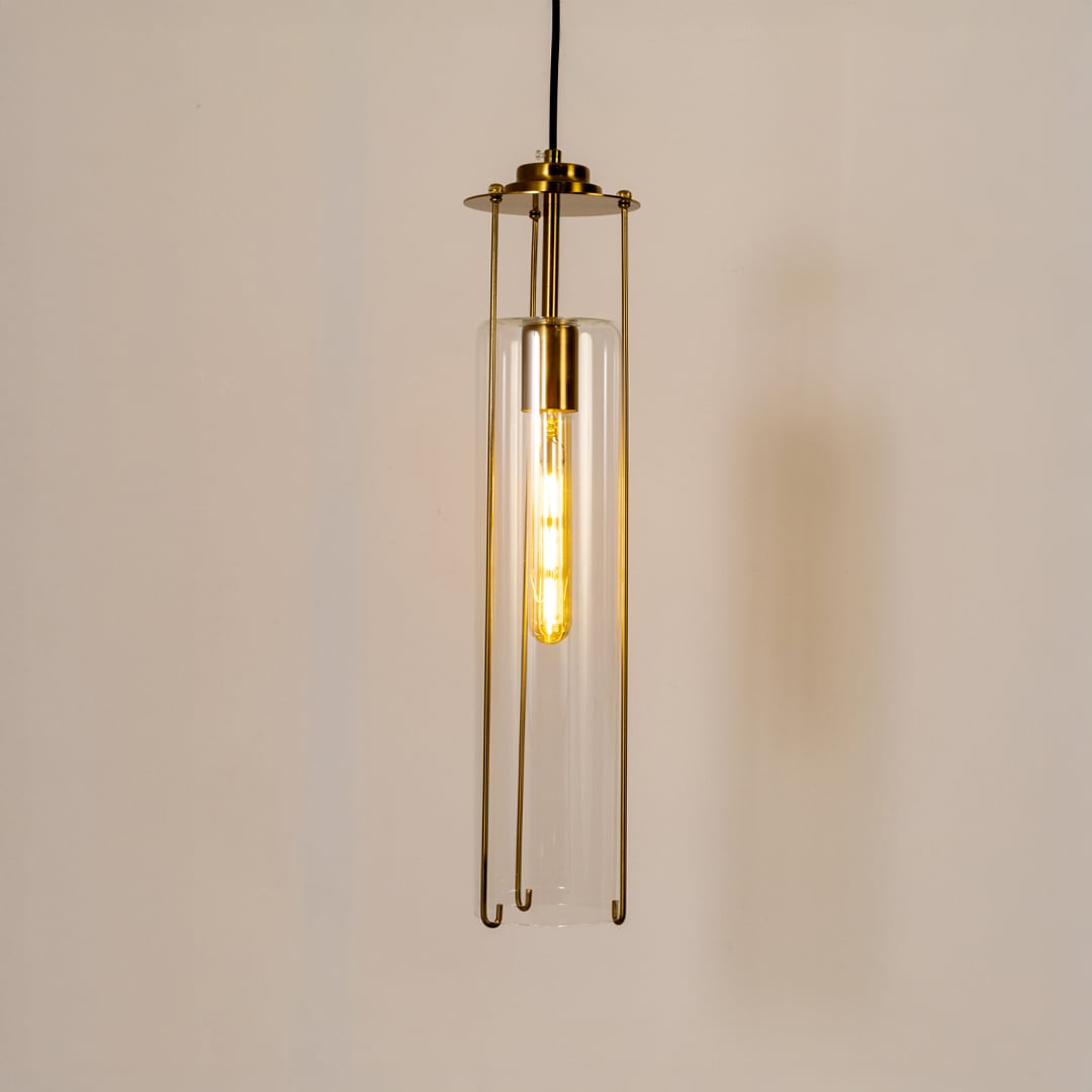 Elegant clear glass pendant light glowing warmly against a clean white background highlighting its slim cylindrical shape and brass accents.