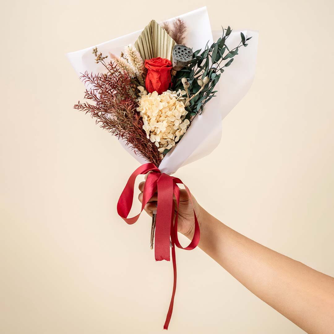 Hand holding a classic red rose dried flower bouquet tied with a red satin ribbon for luxury romantic anniversary gifting.