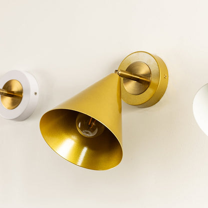 Close-up of a gold cone shaped wall sconce with a visible bulb mounted on a round brass base against a light colored wall.