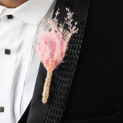 Closeup of boutonniere with soft pink fabric flower and dried grass wrapped in jute on black suit lapel.