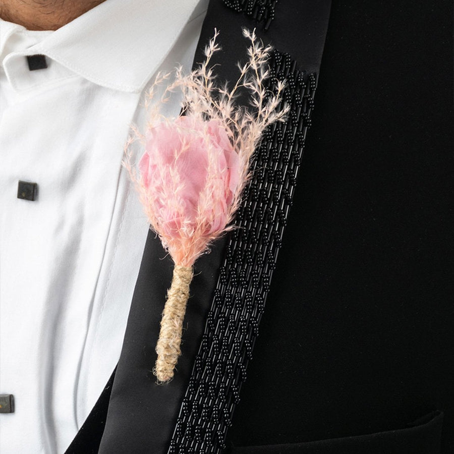 Closeup of boutonniere with soft pink fabric flower and dried grass wrapped in jute on black suit lapel.