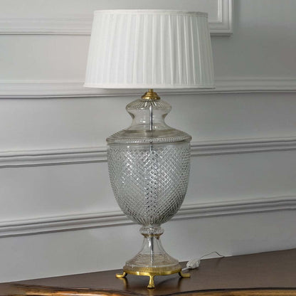 Elegant glass table lamp with brass details and pleated white shade turned off on white background highlighting its exquisite diamond cut pattern.