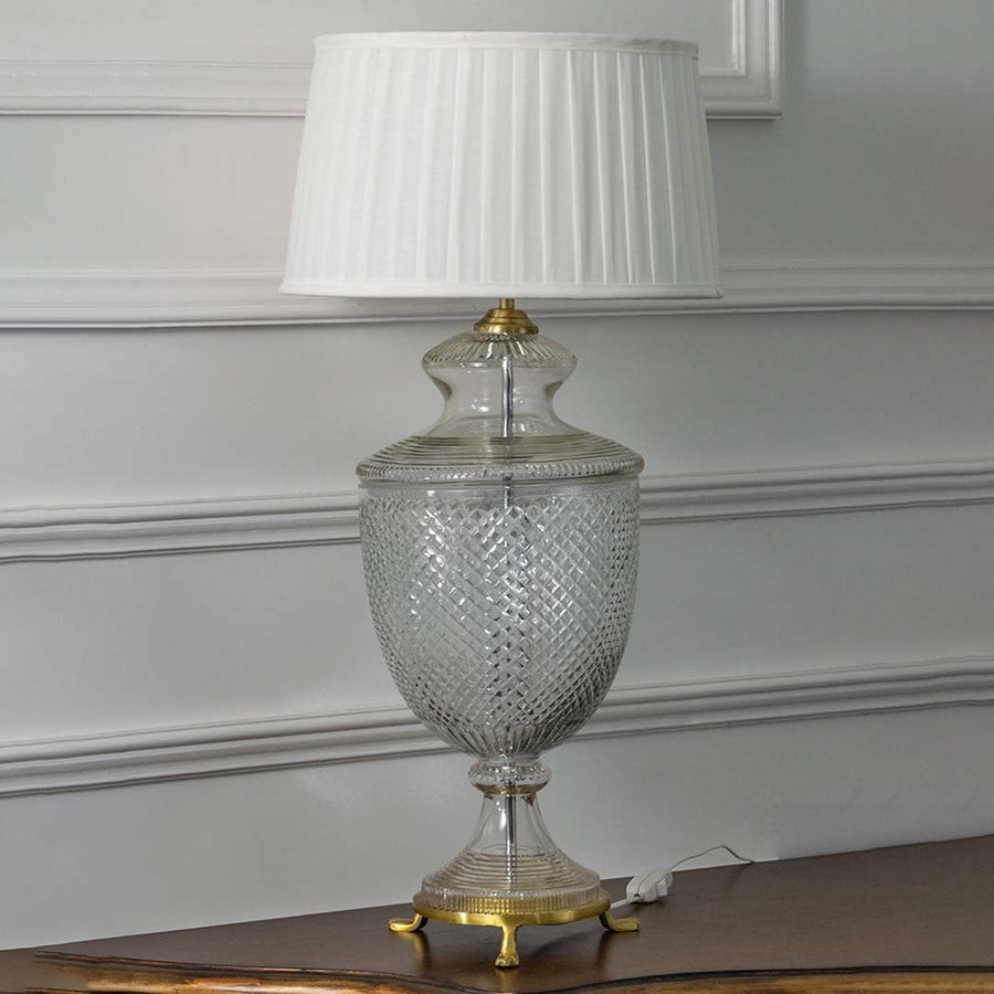 Elegant glass table lamp with brass details and pleated white shade turned off on white background highlighting its exquisite diamond cut pattern.