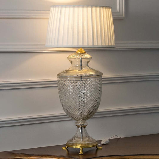 Crystal glass table lamp with brass base and pleated white shade turned on against white background perfect for bedroom or hallway illumination.