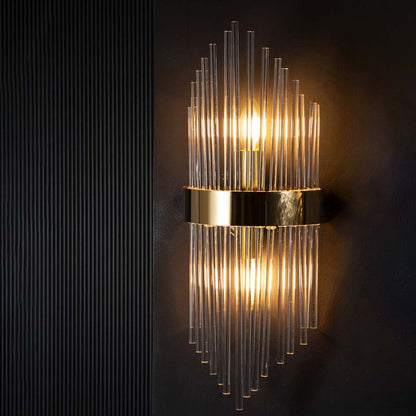 Wall light on a black background with lights on creating a warm radiant glow through the tall glass rods and polished gold metal frame.