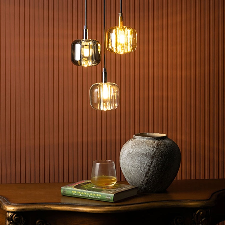 Glass pendant lights in amber and transparent colors suspended above a wooden table with a decorative vase and book in a warm modern living room.