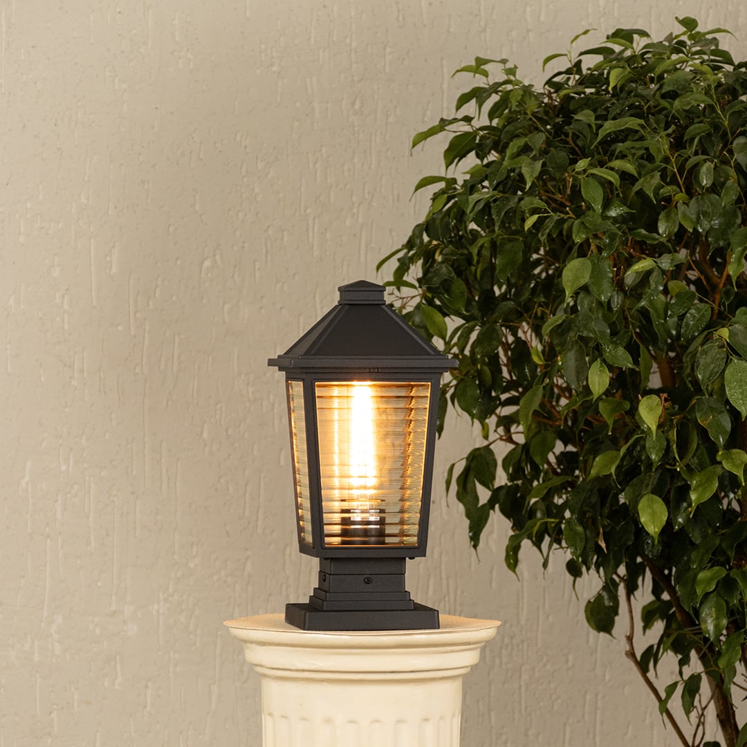 Black outdoor gate light with warm glowing bulb styled beside plants for elegant exterior decor ambiance.