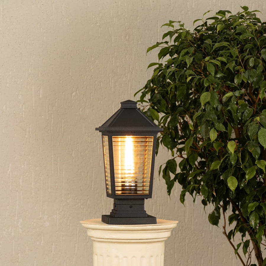 Black outdoor gate light with warm glowing bulb styled beside plants for elegant exterior decor ambiance.