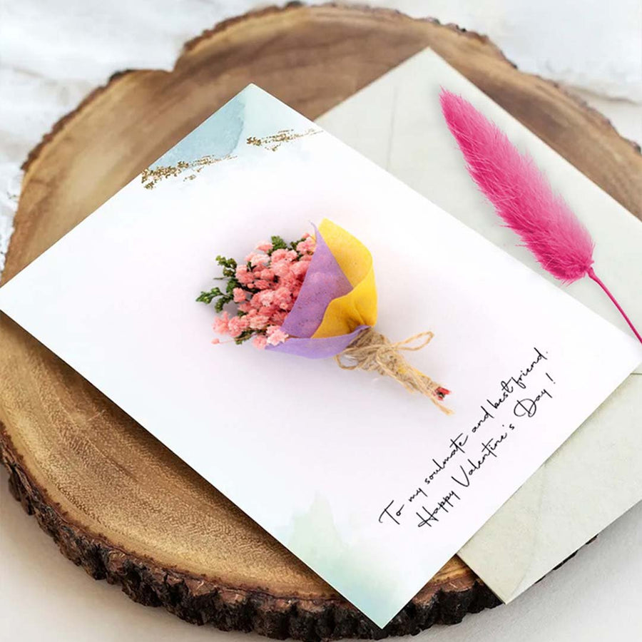 Flat lay of a handmade mini dried flower bouquet greeting card on a wooden log slice next to a bright pink feather quill.