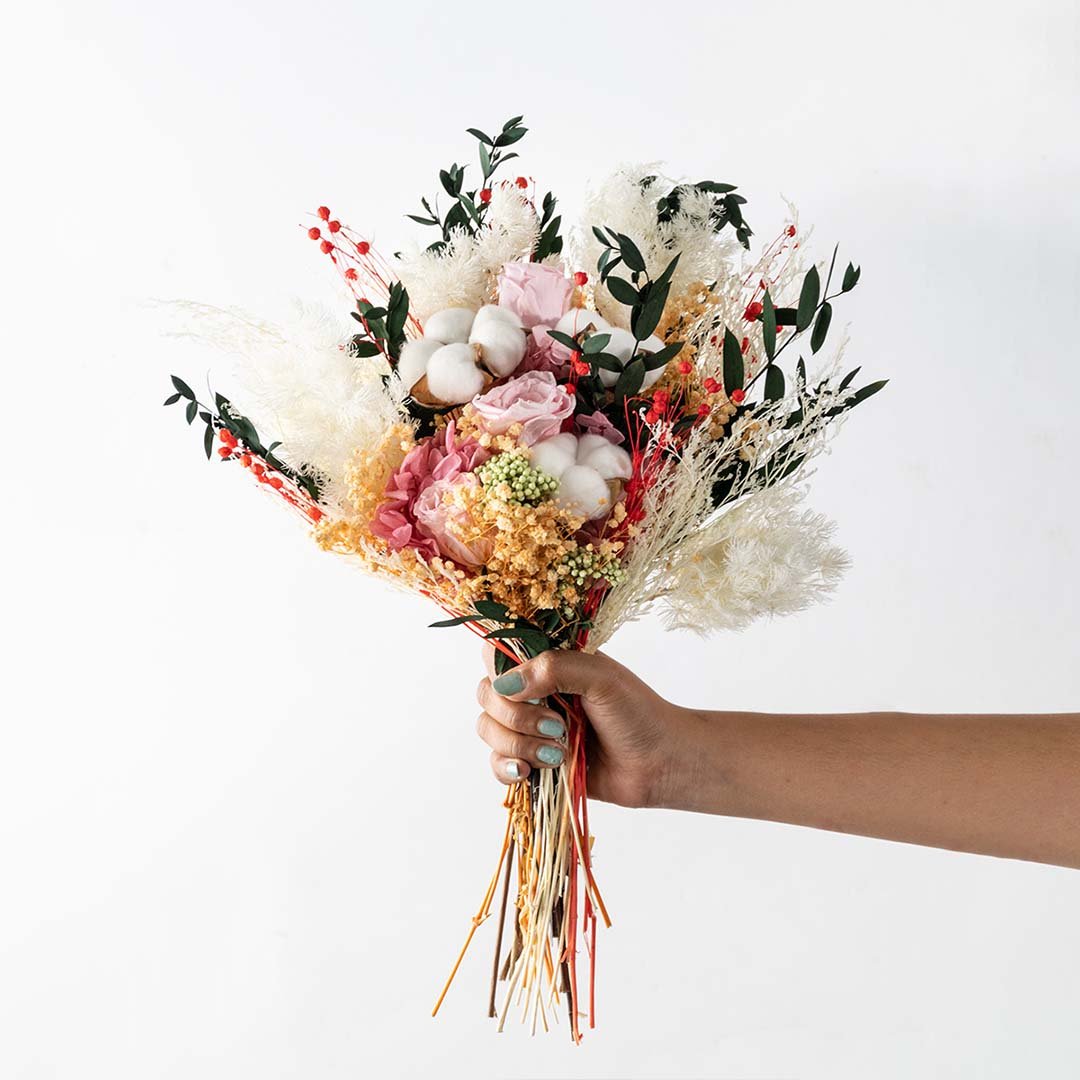 Hand holding a luxury lush dried flower bouquet with pink roses cotton pods and fluffy pampas grass on a white background.
