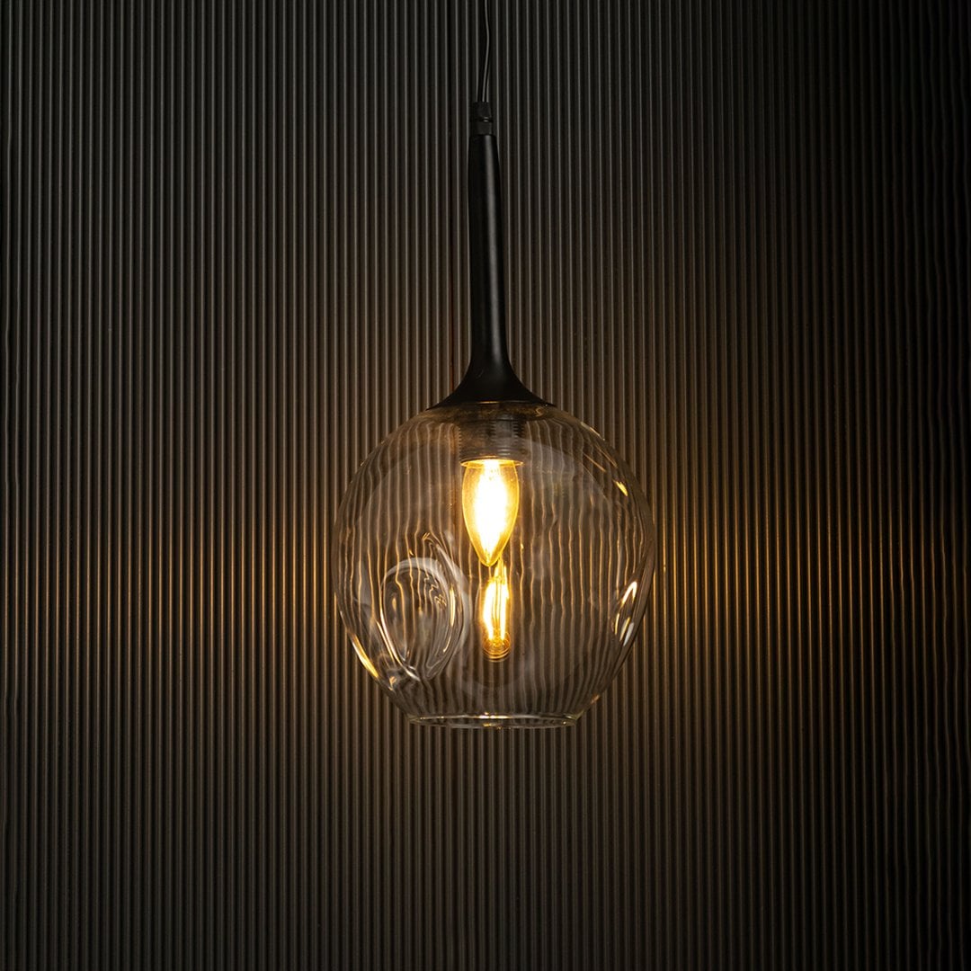 Clear glass pendant light with black fixture glowing warmly against black background.