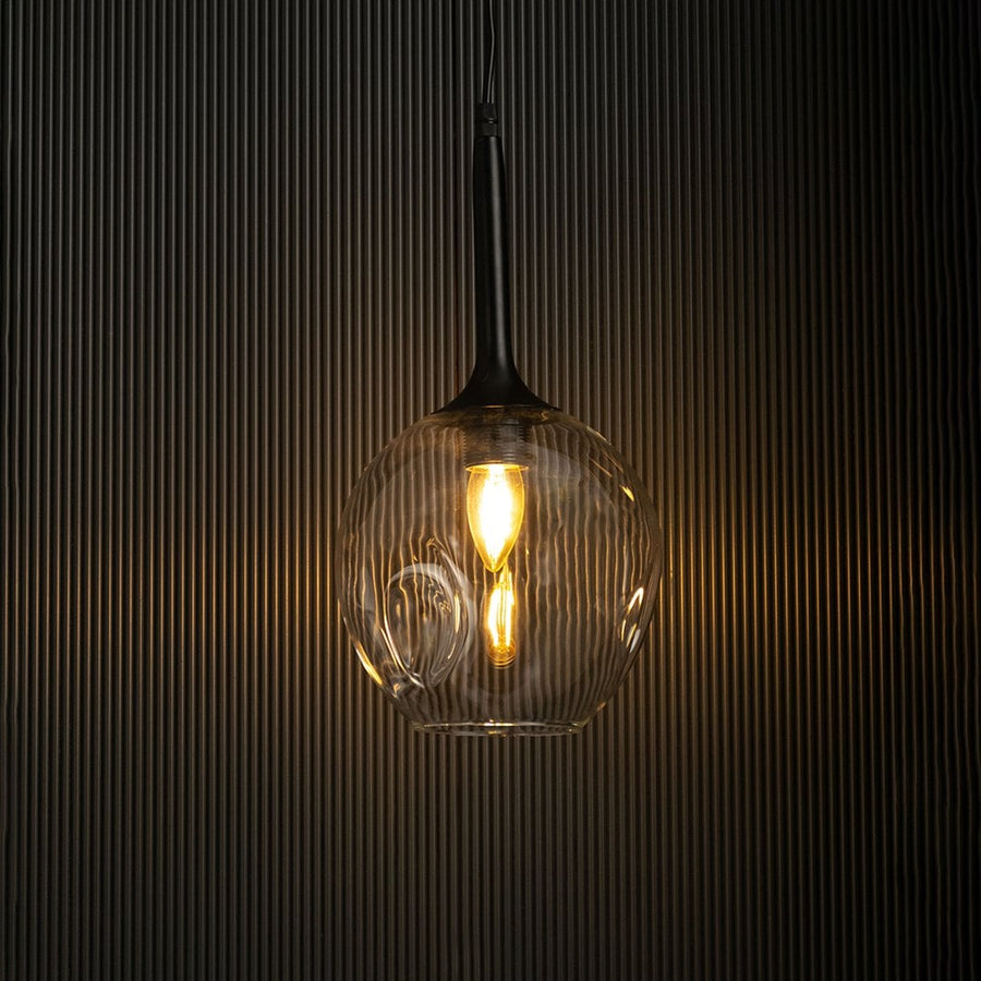 Clear glass pendant light with black fixture glowing warmly against black background.