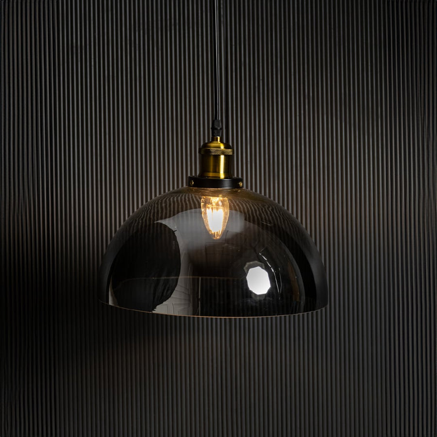 Smokey glass dome pendant light glowing on black background with brass top and black fitting for modern decor.