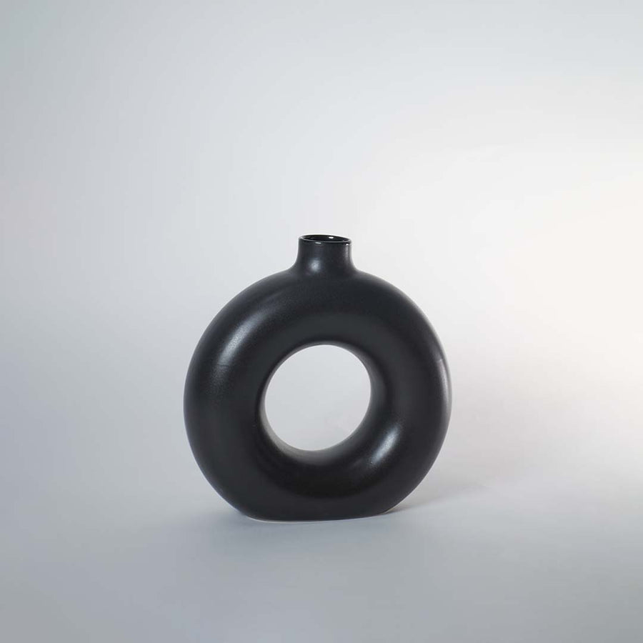 A black circular vase photographed on a white background showing its smooth rounded silhouette and central hollow design that adds a modern artistic touch to decor.
