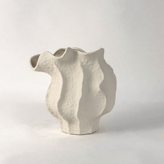 White ceramic sculptural vase with organic flowing shape placed on white surface against neutral background showcasing elegant modern home decor styling.