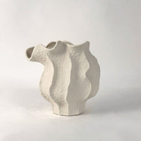 White ceramic sculptural vase with organic flowing shape placed on white surface against neutral background showcasing elegant modern home decor styling.