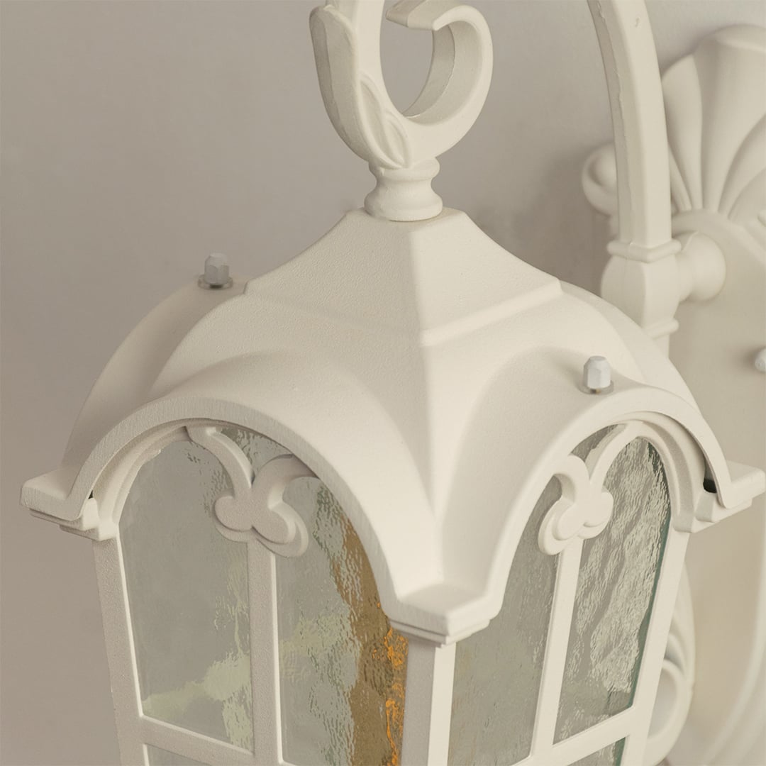 Closeup view of a white outdoor wall light showcasing its arched frame textured glass panels and decorative scroll detailing on the mounting arm.