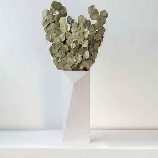 A tall geometric white vase filled with dried rounded eucalyptus style leaves displayed against a soft light background creating natural earthy botanical accents for modern interior décor.