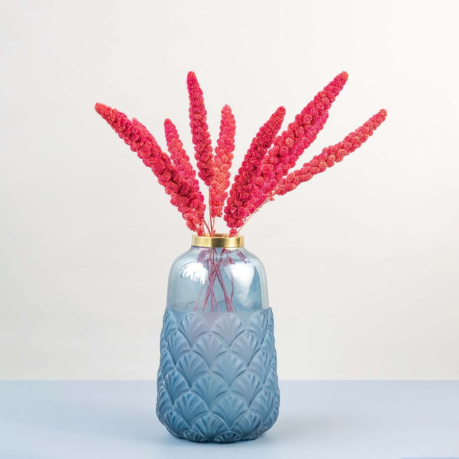 Vibrant pink dried millet stems styled in textured blue glass vase with gold rim creating modern lifestyle home decor.