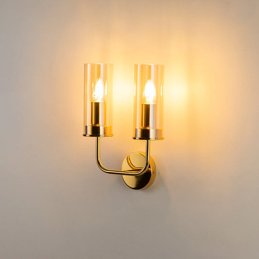 Illuminated double gold wall lamp with clear glass cylinders isolated on white background highlighting bright warm lighting glow.
