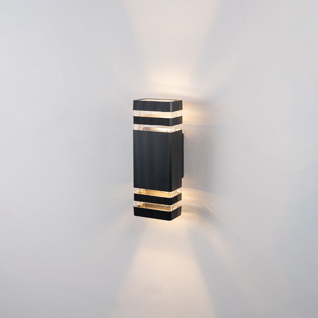 Black metal outdoor wall light glowing on white wall with gold accent bands for elegant exterior decor.