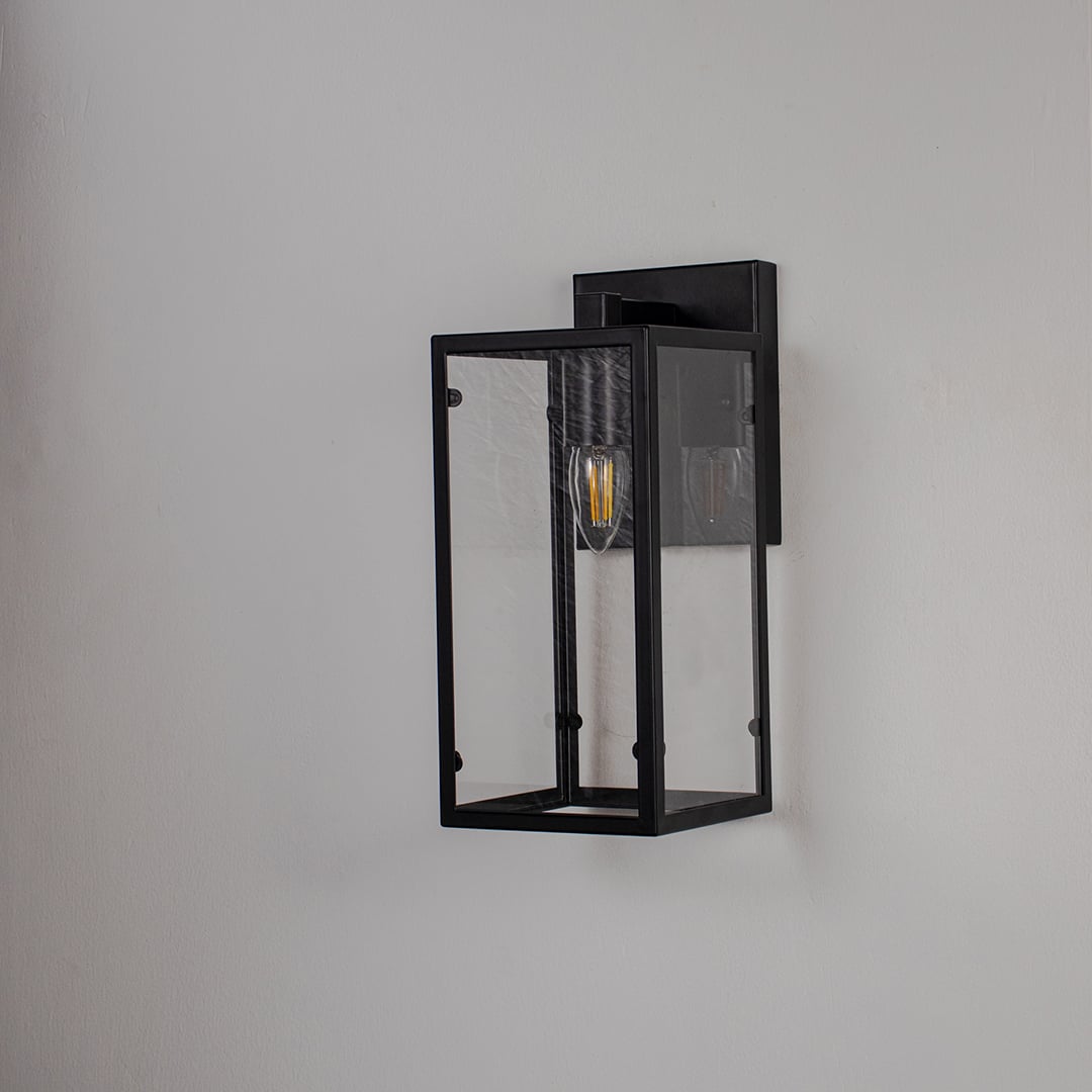 Black rectangular wall lantern with clear glass panels and exposed bulb shown off on white background for modern home decor.