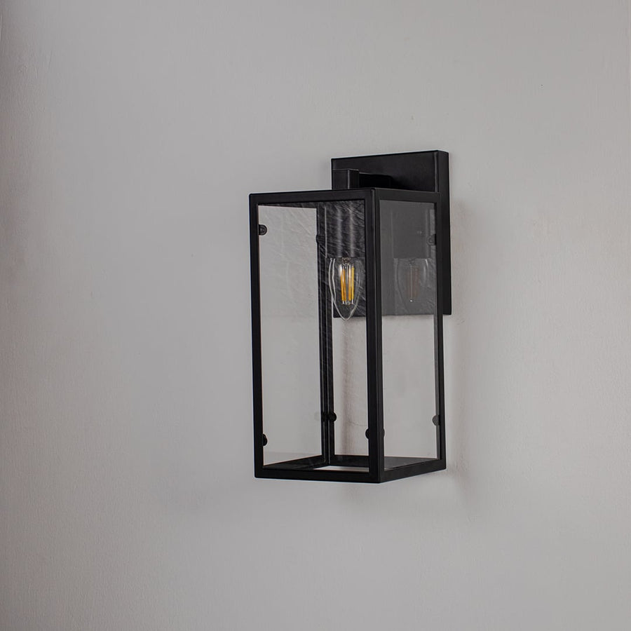 Black rectangular wall lantern with clear glass panels and exposed bulb shown off on white background for modern home decor.