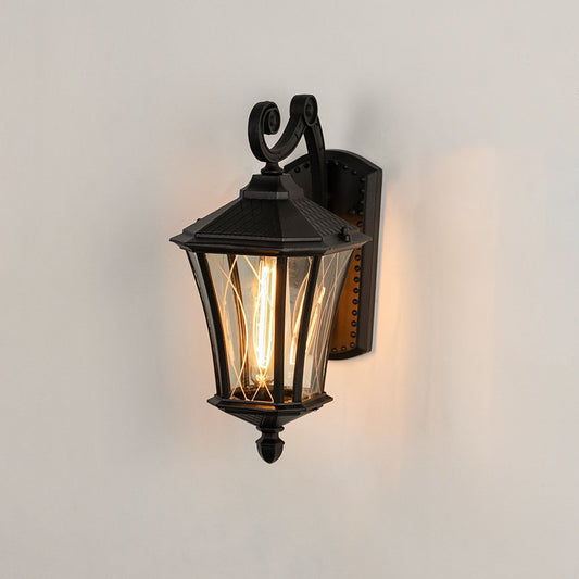 Black metal outdoor wall lantern on white background with clear tapered paneled glass shade for entryway patio decor.