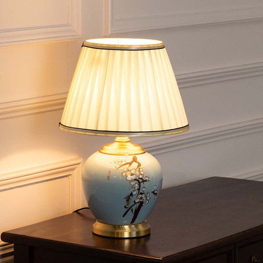 Table lamp glowing softly with blue ceramic base and pleated white shade on a white background for bright decor styling.