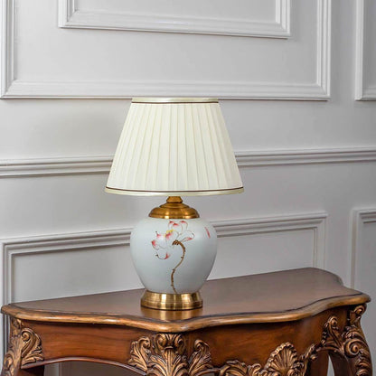 Ceramic table lamp turned off with pleated shade floral ceramic body and golden base on white background.