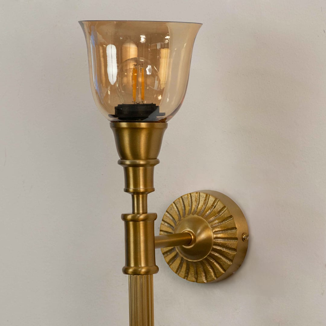 Close up view of brass wall sconce showing ribbed metal arm decorative round backplate and amber glass shade details.